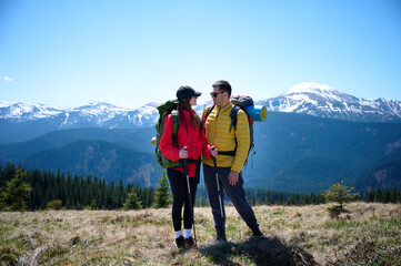 Fototapeta premium Two hikers taking a break on a mountain ridge. Sporty couple with hiking backpacks standing on background of snowy mountains.