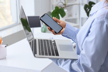 Young woman holding mobile phone with blank screen and modern laptop in office