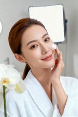 A beautiful, serene woman in a white bathrobe applies a moisturizing face cream to her cheek in soft light, holding the jar and enjoying a skincare routine with a gentle mist and glowing bokeh backgro