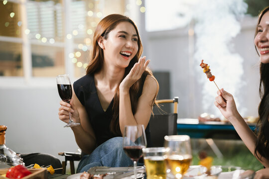 Two young women enjoying grilled food and wine together at an outdoor party, smiling and chatting in a relaxed, friendly atmosphere.