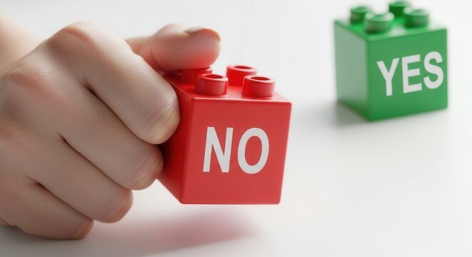 A hand holds a red building block with NO written on it A green building block with YES is blurred in the background on a light surface