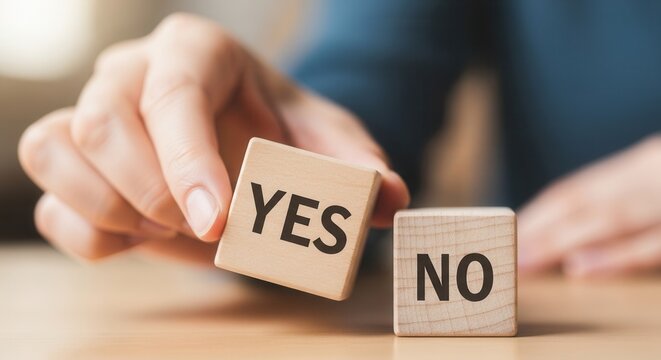 A hand holds a wooden block with YES printed on it positioned above another block labeled NO on a wooden surface