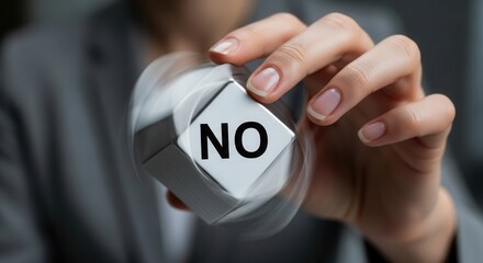 A persons hand holds and spins a silver cube with the word NO in black letters on its white face set against a blurred grey background