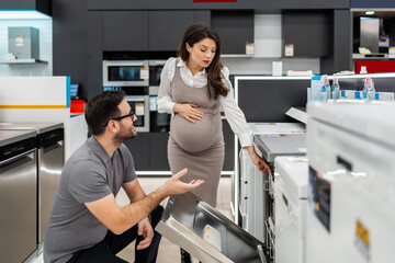 Smiling salesman holding a tablet, discussing and showing dishwashers to a pregnant customer, selecting the best options in a modern home appliance store