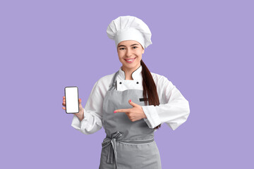 Young female chef pointing at mobile phone on lilac background