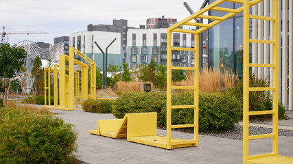 modern outdoor fitness equipment in urban park surrounded by greenery and contemporary architecture, featuring bright yellow structures under cloudy sky, promoting active urban lifestyle