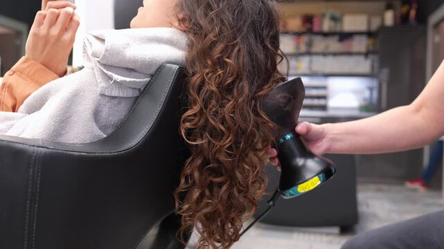 Professional hairstylist drying a client's long, beautiful brown curls with a diffuser hairdryer in a modern beauty salon