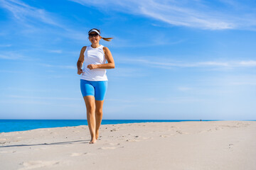 Beautiful mature woman training running on sandy beach in summertime. Physical activity on beach. Front view	