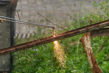Sparks fly as a plasma cutter slices a rusty steel channel outdoors among green grass.