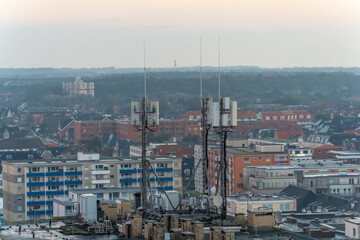 Close-up of antenna masts and satellite dishes on a rooftop. The background shows a city with buildings and distant hills.