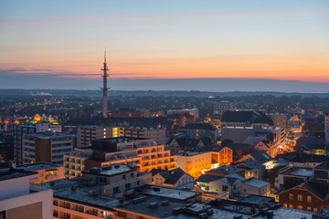Naklejka premium A panoramic view of the city with a tall communications tower at sunset. Warm orange light contrasts with urban rooftops.