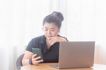Young woman using smartphone while sitting at a desk with a laptop in a bright room during the day