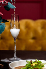 Champagne overflowing in flute glass on elegant table with salad, mustard-yellow couch and red wall in background. Festive moment with foam and sparkling wine splash.