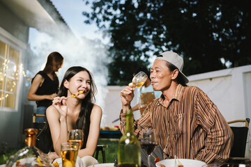 Group of friends enjoying barbecue and wine together in a cozy outdoor party, smiling and chatting in a cheerful atmosphere.