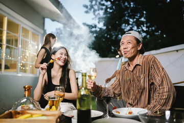 Group of friends enjoying barbecue and wine together in a cozy outdoor party, smiling and chatting in a cheerful atmosphere.