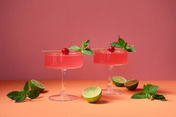 Glasses of tasty Cosmopolitan cocktail garnished cherries and mint on orange table against pink background