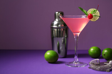 Glass of tasty Cosmopolitan cocktail garnished with cherry and lime on purple background