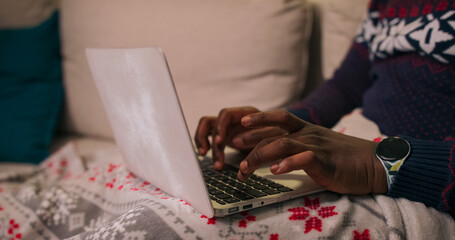 A close-up of the hands of a young man of multi-ethnic background typing on a laptop keyboard. Dressed in a warm holiday sweater with a watch on his wrist and a ball earring, he concentrates on his
