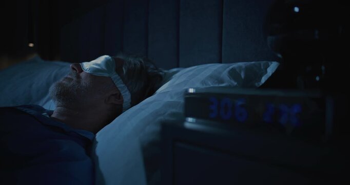 Close Up of White Haired Man Tossing and Turning in Bed at Three AM. Wearing Sleep Mask in Dark Bedroom. Alarm Clock Glows on Bedside Table. Insomnia, Aging, Poor Sleep Hygiene, Mental Health Issues.