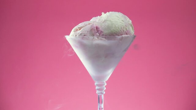 Scoops of vanilla ice cream in a glass goblet slowly rotate against a brightly colored background.