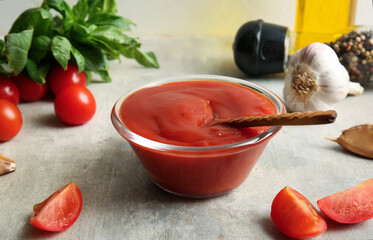 Glass bowl of tasty tomato sauce with garlic and basil leaves on grey background