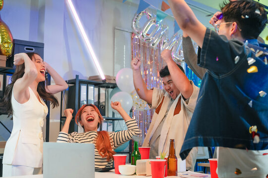 Excited office team celebrates incredible profit growth and victory results announcement together, cheering enthusiastically while monitoring financial performance on screen during triumphant night