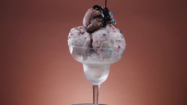 Balls of chocolate ice cream in a glass bowl, drizzled with chocolate fudge, slowly rotate against a brightly colored background.