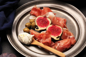 Plate with delicious prosciutto slices, fresh fig, olives and cream cheese on black background, closeup