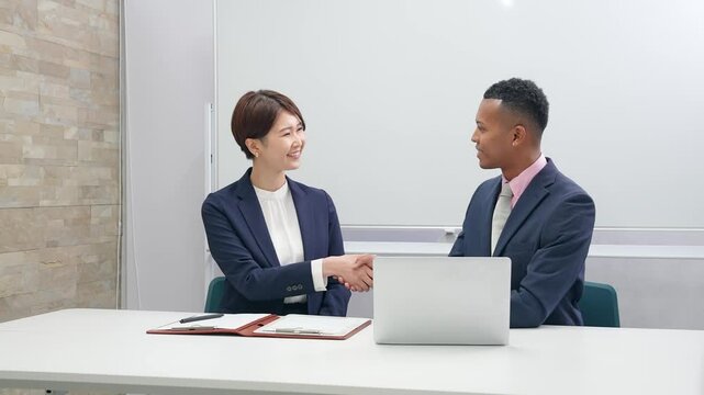 A black male businessman and a Japanese female staff member shaking hands in front of a whiteboard