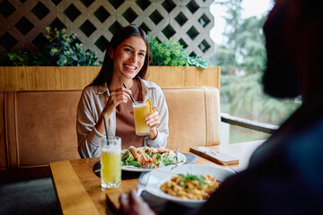 Smiling woman holding a glass with beverage and straw, sitting at a table with food, having lunch with a person