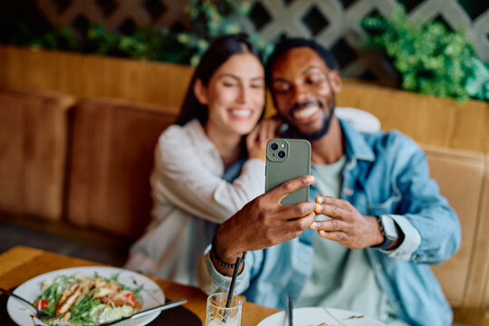 Happy interracial couple enjoying a date, smiling while capturing a selfie at a restaurant table. Modern love and technology connection