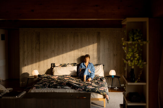 A woman in blue loungewear sits on a bed, sketching or writing on a tablet in warm morning light. The cozy interior with concrete walls and soft fabrics evokes a sense of calm