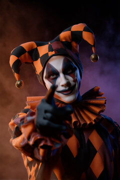 Sinister jester in a harlequin costume pointing at the camera. Scary clown character with evil makeup for Halloween. Theatrical portrait on a dark background