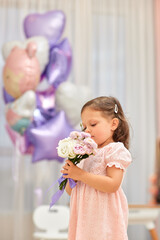 Cute little child girl in pink dress smells flowers on pink purple balloons background. Birthday concept.