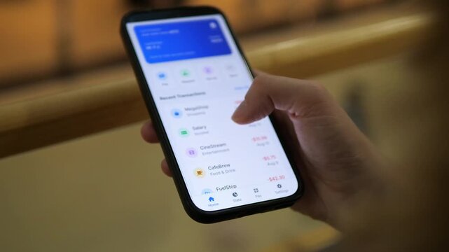 A woman's hands in a public setting scroll through a transaction list in a mobile banking application, locating and selecting a salary deposit entry to view its details. 