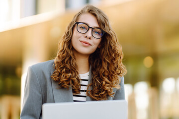Portrait of a stylish woman with a laptop sitting on the steps in front of a modern office building. A beautiful woman in glasses works remotely outdoors. Freelancing concept, weekend.