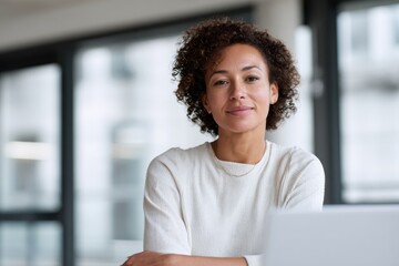 Woman with curly hair wearing a white top smiling while seated at a desk in a modern office environment with natural light and large windows in the background