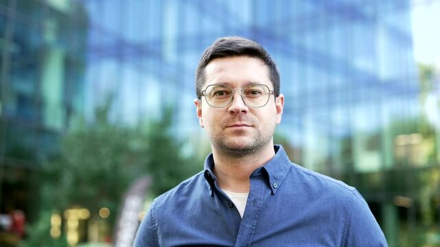 Portrait of a serious businessman standing on street near office building. Headshot of handsome male in blue shirt and glasses looking at camera. Confident business man posing arms outside. Close up