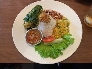 Balinese dish served on white plate featuring Bebek Betutu roast duck, accompanied by amound of rice topped with fried shallots. includes sauteed greens contain grated coconut, roasted peanuts, salad.