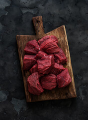 Fresh beef fillet cut into pieces on a rustic wooden board on a dark background, top view