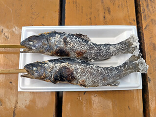 Two grilled ayu fish skewers coated with salt, served on a white tray over a wooden table, representing traditional Japanese street food simplicity.