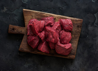 Fresh beef fillet cut into pieces on a rustic wooden board on a dark background, top view