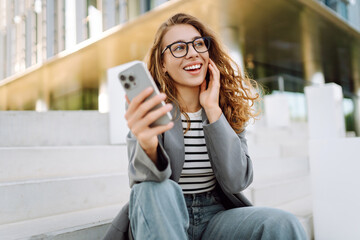 Portrait of stylish woman wearing glasses using phone against backdrop of modern office building....