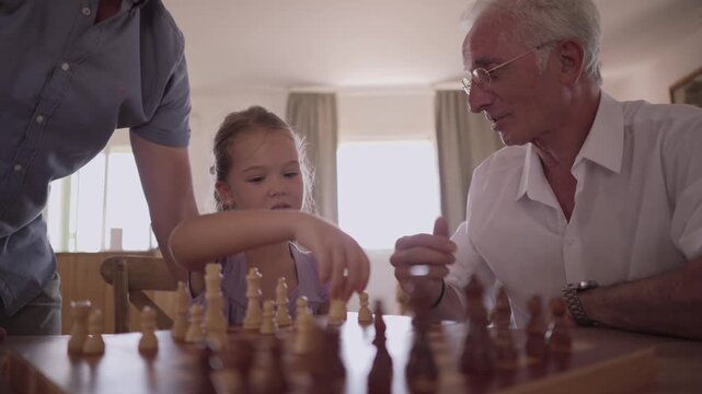 Joyful multi generational family playing a chess board game at home, with the grandfather teaching his granddaughter strategy while the proud father watches and smiles with amusement