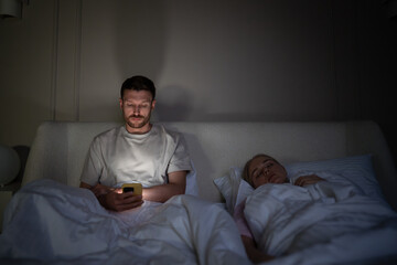 Exhausted man awake alone in bed scrolling phone due to insomnia and digital addiction on late...
