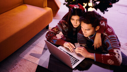 Couple in festive sweaters shopping online at home by a Christmas tree, using a laptop and credit card on a glass table, cozy holiday evening atmosphere.