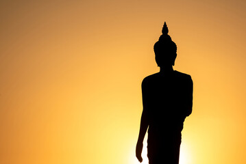 Stand big Buddha Statue in silhouette sun set Light background in park of Thailand temple.Yellow orange light silhouette dark shadow of image Buddha statue stand Buddha tall walk in sun light cloud.
