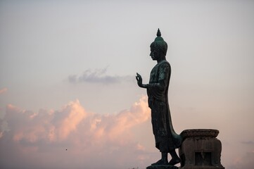 Stand big Buddha Statue in silhouette sun set Light background in park of Thailand temple.Yellow orange light silhouette dark shadow of image Buddha statue stand Buddha tall walk in sun light cloud.