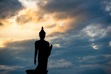 Stand big Buddha Statue in silhouette sun set Light background in park of Thailand temple.Yellow orange light silhouette dark shadow of image Buddha statue stand Buddha tall walk in sun light cloud.