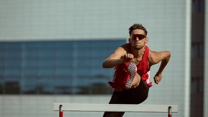 Focused hurdler mid-jump during track competition wearing red uniform. Concept of determination,...